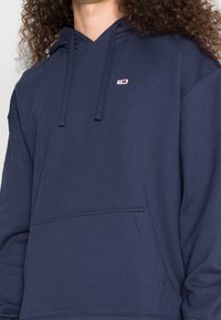 Mörkblå huvtröja i bomullsblandning, med en ficka fram, dragsko på huven och liten logotyp på bröstet. Mjuk, texturerad tyg.