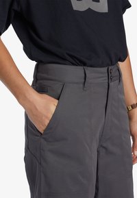 Grå cargo byxor med en slät yta, med två framfickor och en knappstängning vid midjan. Kombineras med en svart topp.