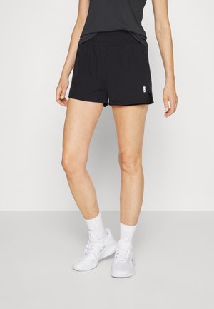 Kobieta ubrana w czarne sportowe szorty, białe skarpetki i białe sneakersy stojąca na tle jednolitego jasnego tła.