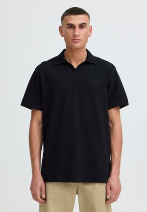 Joven con cabello corto que lleva una camisa polo negra de manga corta y pantalones beige, de pie contra un fondo claro y liso.