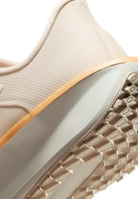 Zapatilla deportiva de color beige claro con parte superior de malla texturizada, banda de acento naranja y suela de goma con un diseño de banda para tracción.