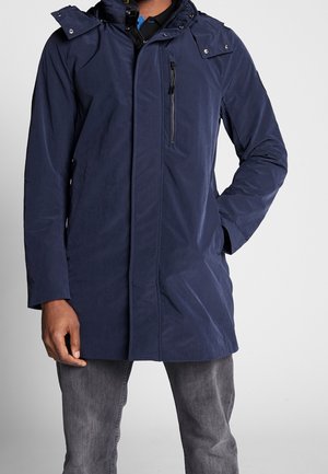 Homme portant un manteau d'hiver bleu foncé à capuche avec une poche zippée devant, une couche intérieure noire et un jean gris sur un fond uni.