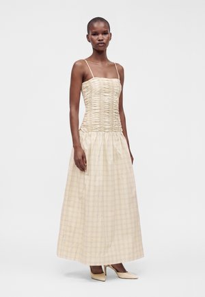 Mujer alta con pelo corto que lleva un vestido largo beige con tirantes finos y un corpiño ajustado con fruncidos, combinado con zapatos de tacón altos beige.