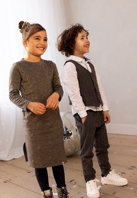 Två barn står och ler. En bär en glittrig grå klänning och svarta leggings; den andra har på sig en grå väst över en vit skjorta och grå byxor.