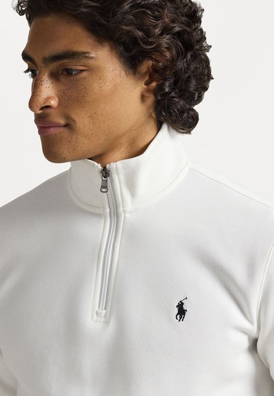 Uomo con capelli ricci che indossa un pullover bianco a zip parziale con un logo di un giocatore di polo nero sul petto, che guarda leggermente di lato.