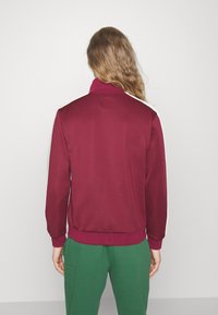 Bordo krāsas zip-up jaka ar augstu apkakli un baltām sānu svītrām, izgatavota no gludas auduma. Izcili riboti roku gali un apakšmala, savienota ar zaļām biksēm.