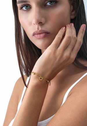 Bracciale in oro con ciondolo a forma di cuore e perline decorative, caratterizzato da un design delicato. Indossato su un polso su uno sfondo minimalista.