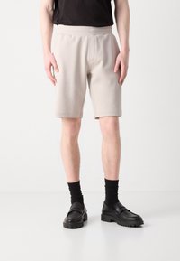 Shorts de sport beiges avec un waistband élastique et des détails de couture, assortis à des chaussettes noires jusqu'au mollet et des chaussures noires à enfiler avec une semelle texturée.