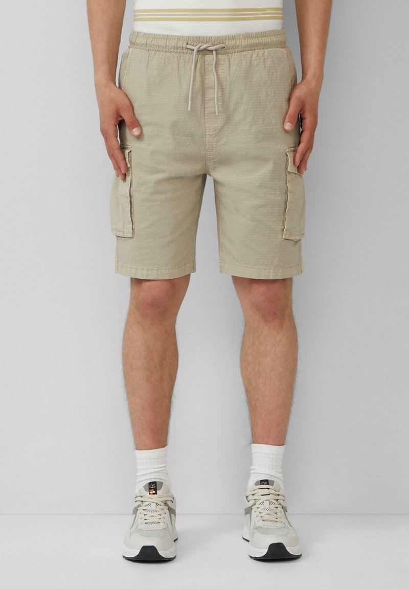 Beige Cargo-Shorts mit einem elastischen Bund und Kordelzug, ausgestattet mit seitlichen Taschen und einem strukturierten Stoff. Getragen mit hellen Sneakers.