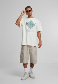 Kremfarget grafisk t-skjorte med teal trykk; beige cargo shorts med sidelommer; hvite sokker; lyse joggesko; person med solbriller.