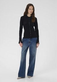 Žena stojaca v čierno zapnutej košeli s dlhým rukávom, v širokých modrých džínsach a špičatých sivých topánkach na pozadí s obyčajným bielym pozadím.