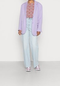 Blazer oversize viola sopra un top a fantasia floreale rossa; jeans a gamba larga blu chiaro con orli sfrangiati; sneakers rosa e bianche.
