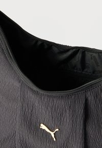 Μαύρη υφασμάτινη τσάντα Puma με χρυσό λογότυπο, ανοιχτό φερμουάρ που αποκαλύπτει μαύρη εσωτερική επένδυση με διακριτική σήμανση Puma.