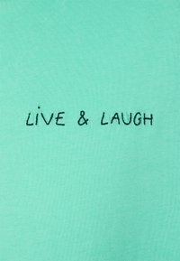 Tyrkysová látka s hladkým povrchem, na které je černým vyšitým textem v neformálním písmu napsáno "ŽIJ A SMÍJ SE".