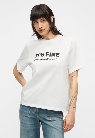 Λευκό βαμβακερό T-shirt με κοντά μανίκια. Στο μπροστινό μέρος υπάρχει έντονο μαύρο κείμενο: «ΕΙΝΑΙ ΕΝΤΑΞΕΙ» και «απλά βάλε ένα φίλτρο πάνω του». Κλασική εφαρμογή.