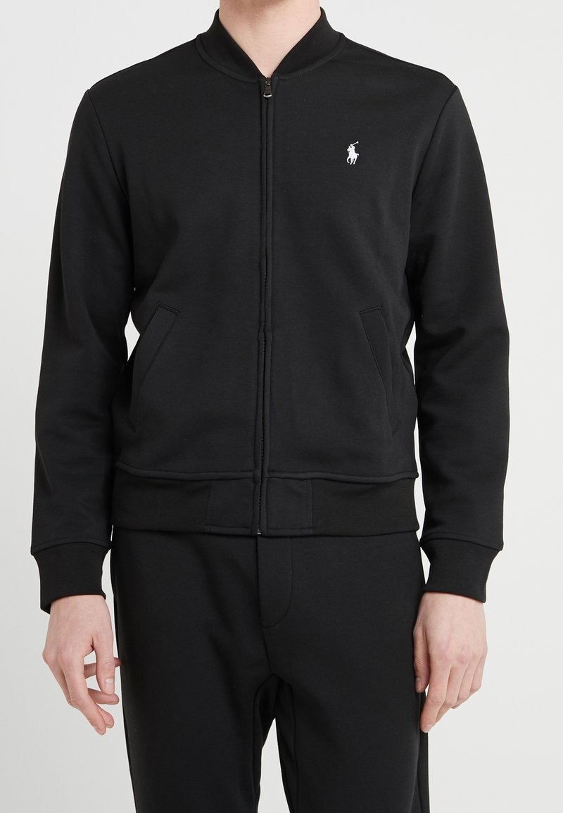 Homme portant une veste noire zippée avec un petit logo blanc de joueur de polo sur la poitrine et un pantalon noir assorti, debout devant un fond uni.