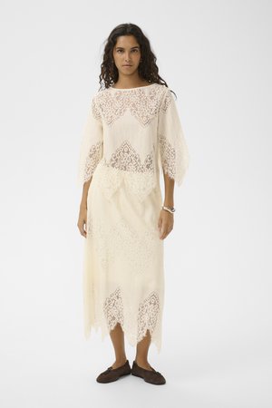 Frau mit lockigem Haar, die ein cremefarbenes Spitzenkleid mit gewellten Kanten und dunkelbraunen Ballerinas trägt, vor einem einfarbigen Hintergrund stehend.