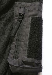 Svart fleece-stoff har en sidelomme med glidelås og en borrelåspatch. Teksturen er myk, med forsterkede sømmer.