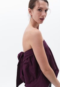 Bordowy top typu off-the-shoulder o luźnym fasonie, z dużą kokardą na plecach i teksturowanym bawełnianym materiałem.