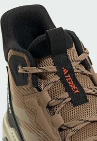 Hnedá turistická obuv s vrchnou časťou z sieťoviny a syntetického materiálu, čierny golier, kontrastné šnúrky a logo nášivka. Obsahuje štruktúrované detaily a vystužené oblasti.