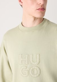 Világoszöld pulóver, bordás kerek nyakkal, a középen dombornyomott "HUGO" márkázással, sima textúrával és laza szabással.