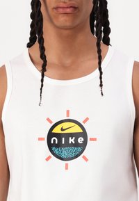 Balta kokvilnas bezmugurkaula kreklā ir apļveida Nike logo ar dzeltenu pusi sauli, melnu tekstu un zilu rakstainu apakšdaļu. Logo ir apņemts ar spilgtiem akcentiem.