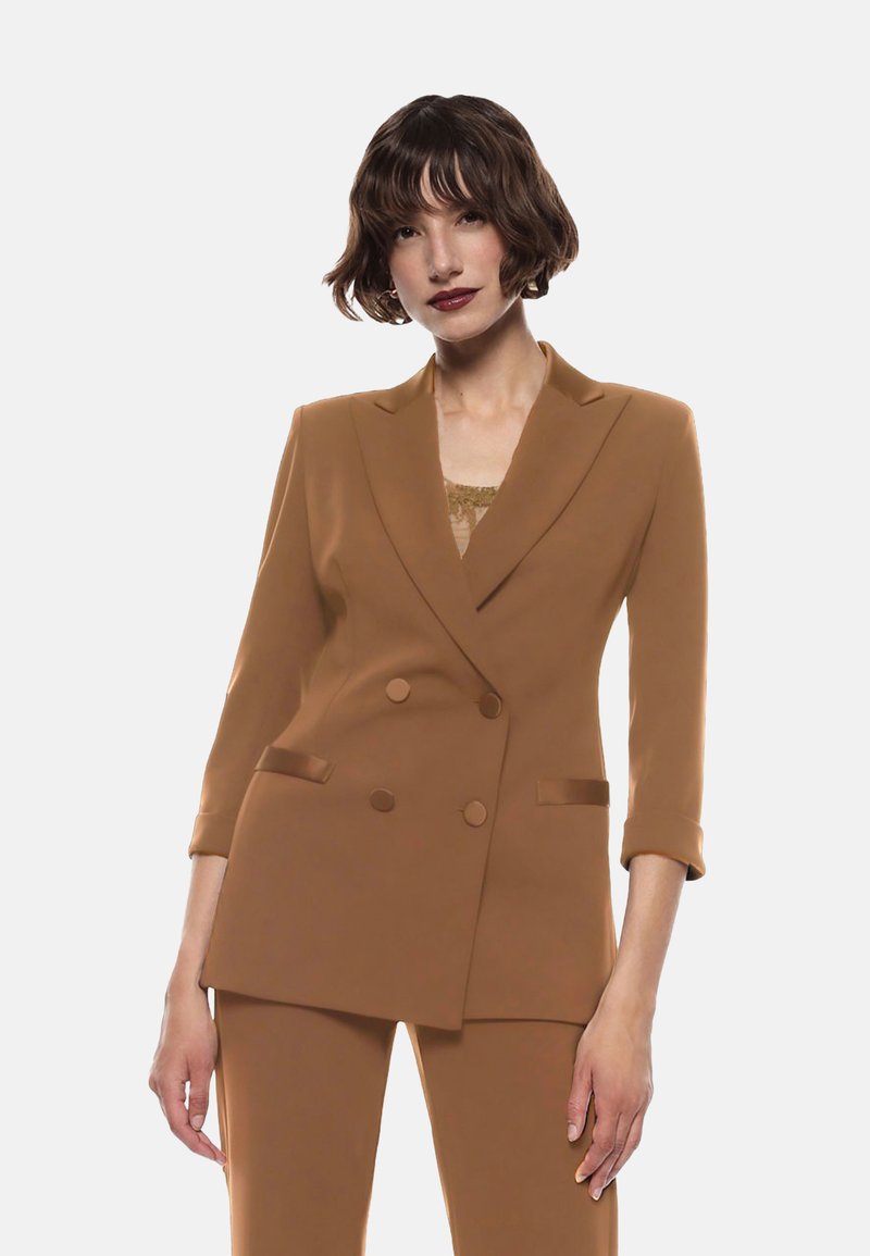 Blazer marrone doppiopetto con maniche a tre quarti, texture liscia e tasche laterali. Indossato sopra un top in pizzo chiaro.