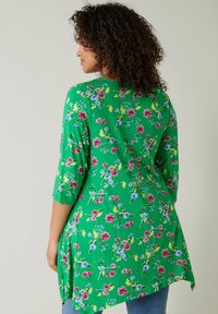 Grünes Blüten-Tunikakleid mit 3/4-Ärmeln, das pinke und blaue Blumen zeigt und ein asymmetrisches Saumdesign hat. Weicher, leichter Stoff mit glatter Textur.