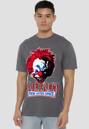Junger Mann trägt ein graues T-Shirt mit einem gruselig aussehenden Clownkopf mit roten Haaren und dem Text "Killer Klowns from Outer Space."