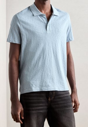 Mann trägt ein hellblaues kurzärmeliges Polohemd und dunkle Jeans, steht mit entspannten Armen seitlich vor einem einfarbigen Hintergrund.