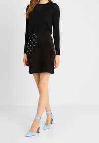 Černý dlouhý rukáv top s průhlednými akcenty, spárovaný s tmavě hnědou semišovou sukní se světle modrými výšivkami hvězd a modrými botami na blokovém podpatku.