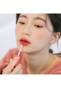 Gloss à lèvres dans un tube transparent avec un applicateur effilé, appliqué sur des lèvres pulpeuses. Les lèvres sont d'une teinte rouge brillante, avec un arrière-plan flou.