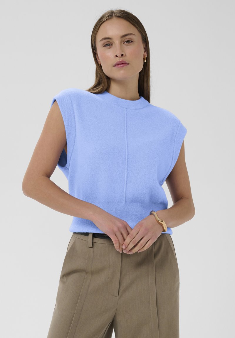 Nuori nainen, joka on pukeutunut olkaimettomaan vaaleansiniseen toppiin, beigeihin korkea vyötäröisiin housuihin, kultaketjuun ja pieniin rengaskorvakoruihin, seisoo yksiväristä taustaa vasten.