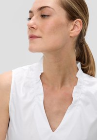 Biała bluzka z falbaniastym dekoltem i bez rękawów. Gładki materiał, prosta sylwetka i subtelne detale wokół kołnierza.