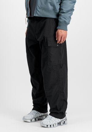 Man wearing black cargo pants with side flap pockets, a blue jacket, and silver athletic shoes with visible air cushioning soles.