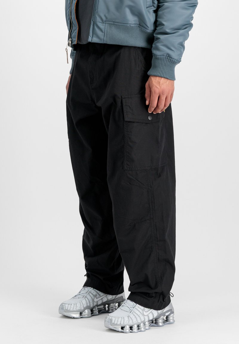 Hombre que lleva pantalones cargo negros con bolsillos laterales con solapa, una chaqueta azul y zapatillas deportivas plateadas con suelas de amortiguación de aire visibles.