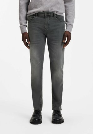 Person iført grå slim fit jeans, sorte snørebåde og en lysegrå sweater, der står mod en hvid baggrund.