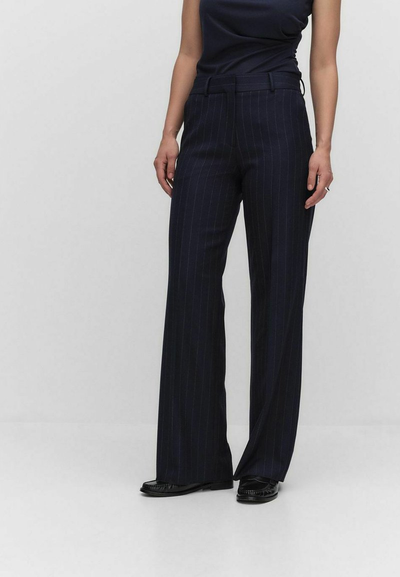 Pantalons rayés bleu marine à jambes larges, avec une texture lisse, des passants de ceinture et une coupe ajustée. Portés avec des chaussures noires.