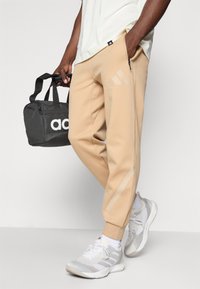 Calças de treino bege com pernas ajustadas, apresentando riscas discretas da marca e fechos de correr, combinadas com um saco de desporto preto e ténis cinza claro.