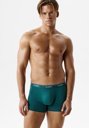 Młody muskularny mężczyzna o jasnobrązowych włosach, ubrany w turkusowe bokserki z napisem "ATLANTIC" na pasie, stojący na jednolitym białym tle.