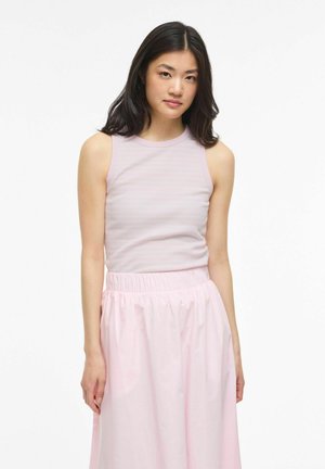 Noor naine, kellel on pikad tumedad juuksed, kannab varrukateta heleroosat topi ja kõrge vöökohaga heleroosat seelikut, seisab valge tausta ees, näoga otse vaataja poole.