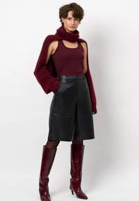 Vrouw in een bordeauxrode tanktop, met een bijpassende gebreide trui over haar schouders, zwarte leren shorts tot de knie en bordeauxrode laarzen tot de knie.