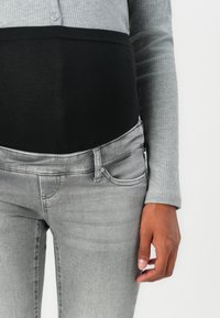 Bluză de maternitate din material ribbed gri deschis, combinată cu o bandă elastică neagră, purtată cu blugi gri cu croială slim și buzunare frontale.