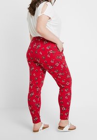 Žena v červených květovaných kalhotách, bílém tričku s krátkým rukávem a výřízky na ramenech a bílých sandálech, stojící před jednobarevným pozadím.