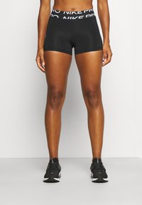 Černé atletické šortky s širokým páskem s logem, přiléhavým střihem a hladkou texturou, v kombinaci s černými atletickými botami.