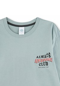 Hellgrüner Sweatshirt mit rundem Halsausschnitt. Mit einem bedruckten Design: "ALWAYS AWESOME CLUB" in Schwarz und Pink, mit einem Etikett am Hals.