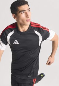 Athlete wearing black Adidas sportswear with red stripes prepares to sprint or change direction against a plain background.