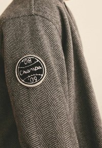 Gros plan sur la manche d'une veste en chevrons grise avec un patch rond noir et blanc indiquant "08 Champs '09" cousu sur le haut du bras.