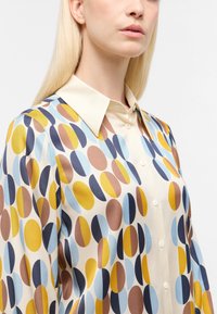 Blusa trasparente color crema con motivi ovali colorati in blu, giallo, marrone e blu navy. Presenta un colletto a punta e chiusura frontale con bottoni.