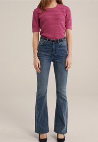 Fuchsiový svetr s texturou a krátkými rukávy, doplněný tmavě modrými rozšířenými džínami a černým páskem, dokončený černými podpatky.
